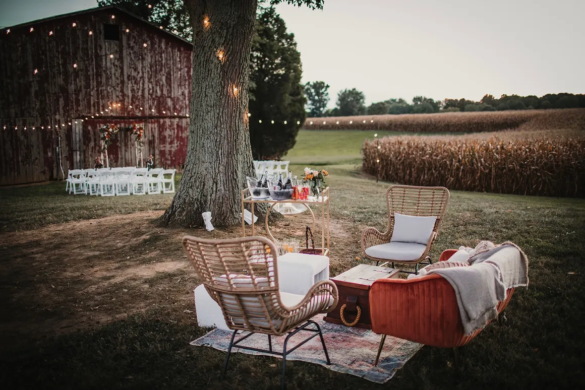The venue at dusk — lounge seating area with wicker chairs, the old barn, café lights, and corn fields