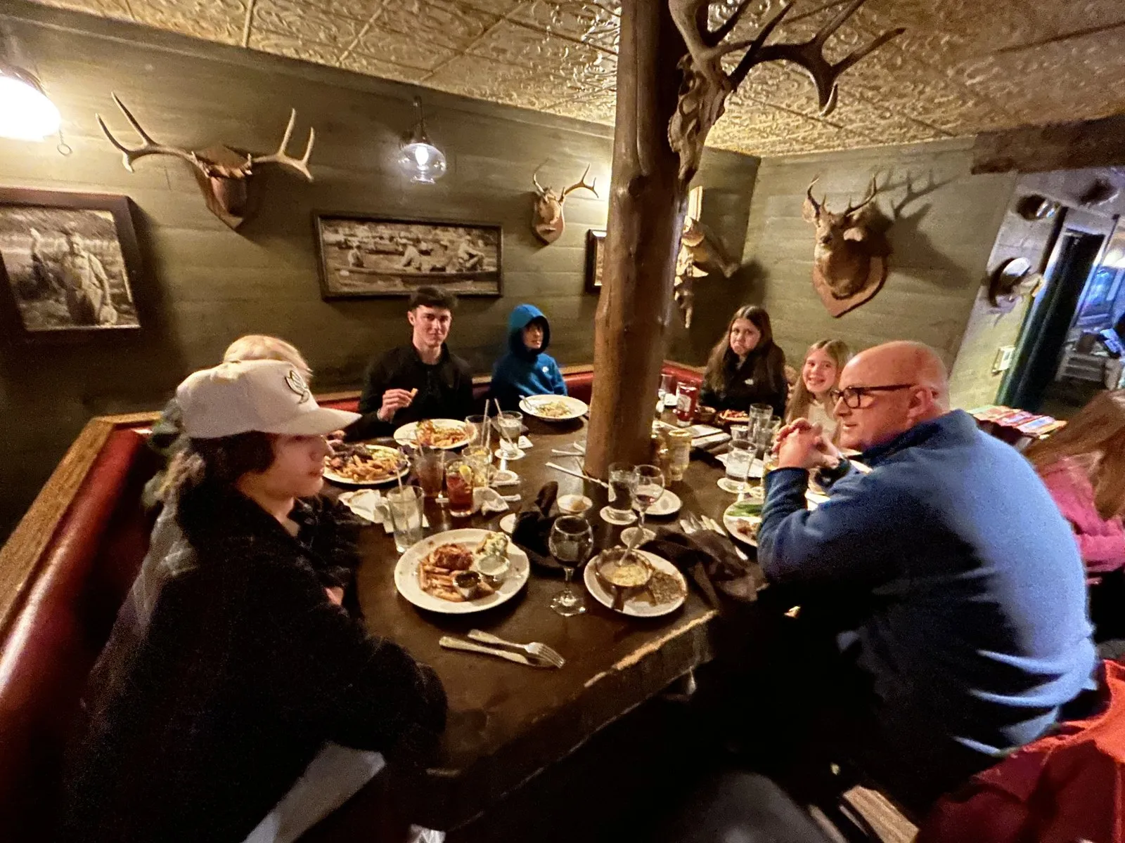 Family at a big table in the Devil’s Pool basement with antler mounts, a tin ceiling and a tree trunk through the middle