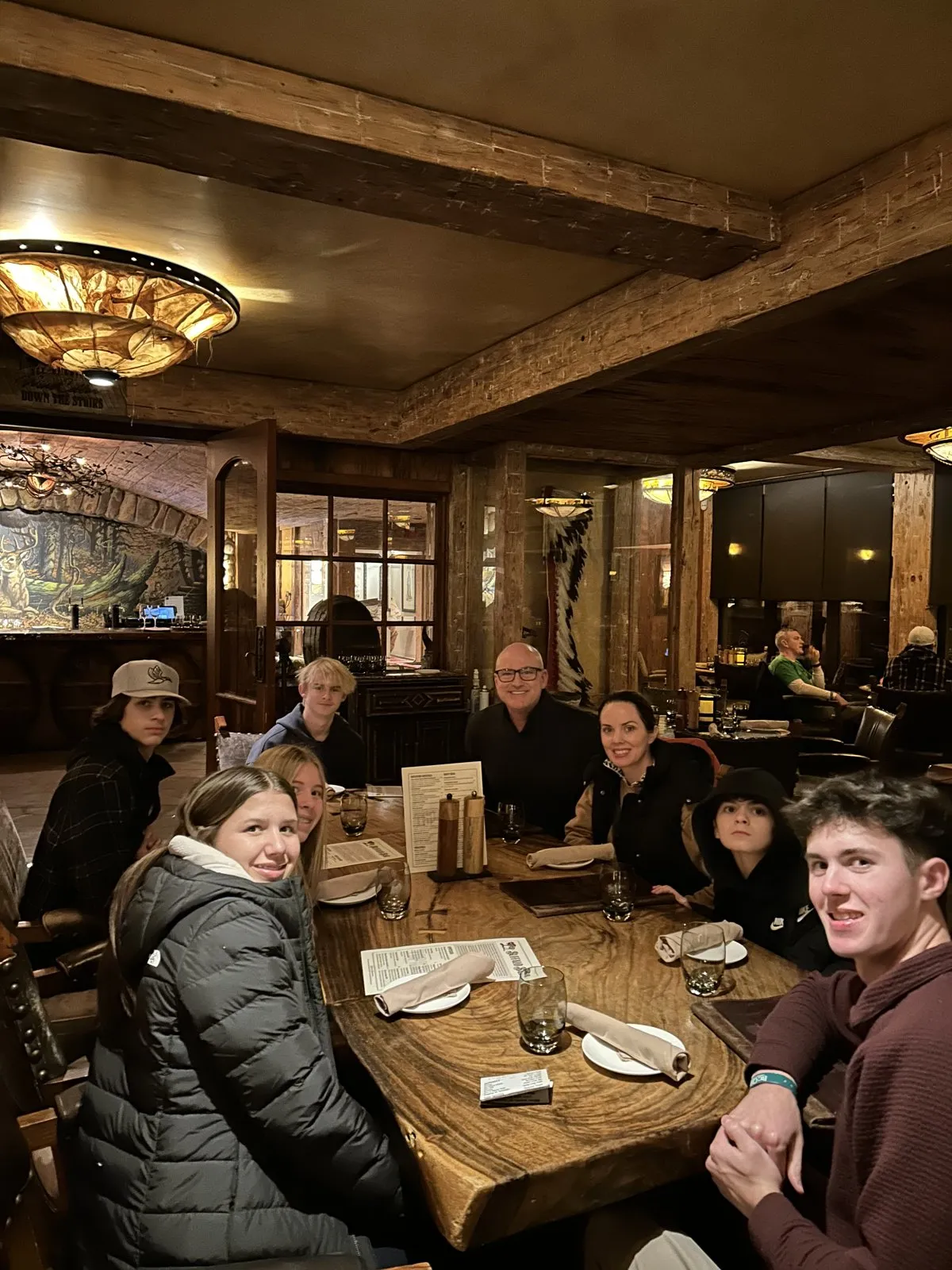 Family around a long live-edge wood table under timber beams and amber chandeliers in the End of the Trail Wine Cellar