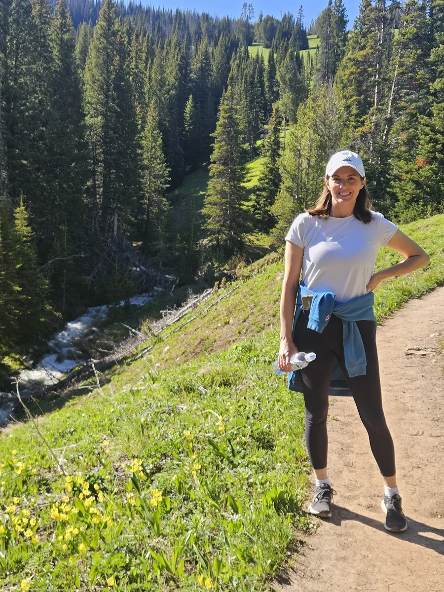 Woman standing on a dirt hiking trail above a creek with wildflowers and pine forest near Big Sky, Montana