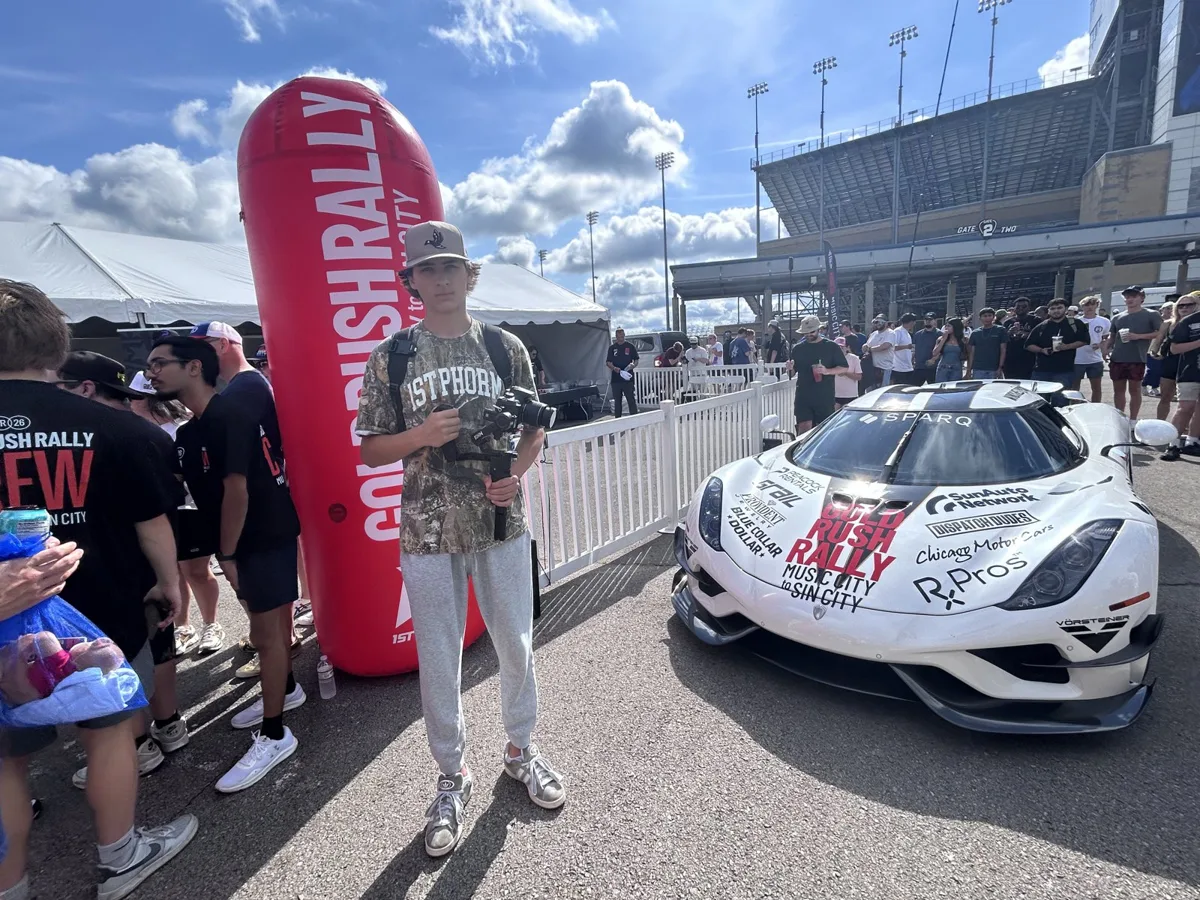 A teen with a camera gimbal next to a wrapped white Koenigsegg at the goldRush Rally