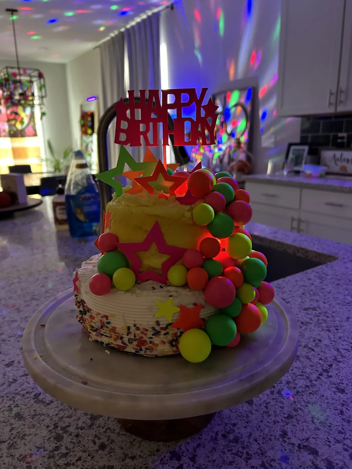 Two stacked sprinkle cakes covered in neon UV cake balls and star toppers with a Happy Birthday topper