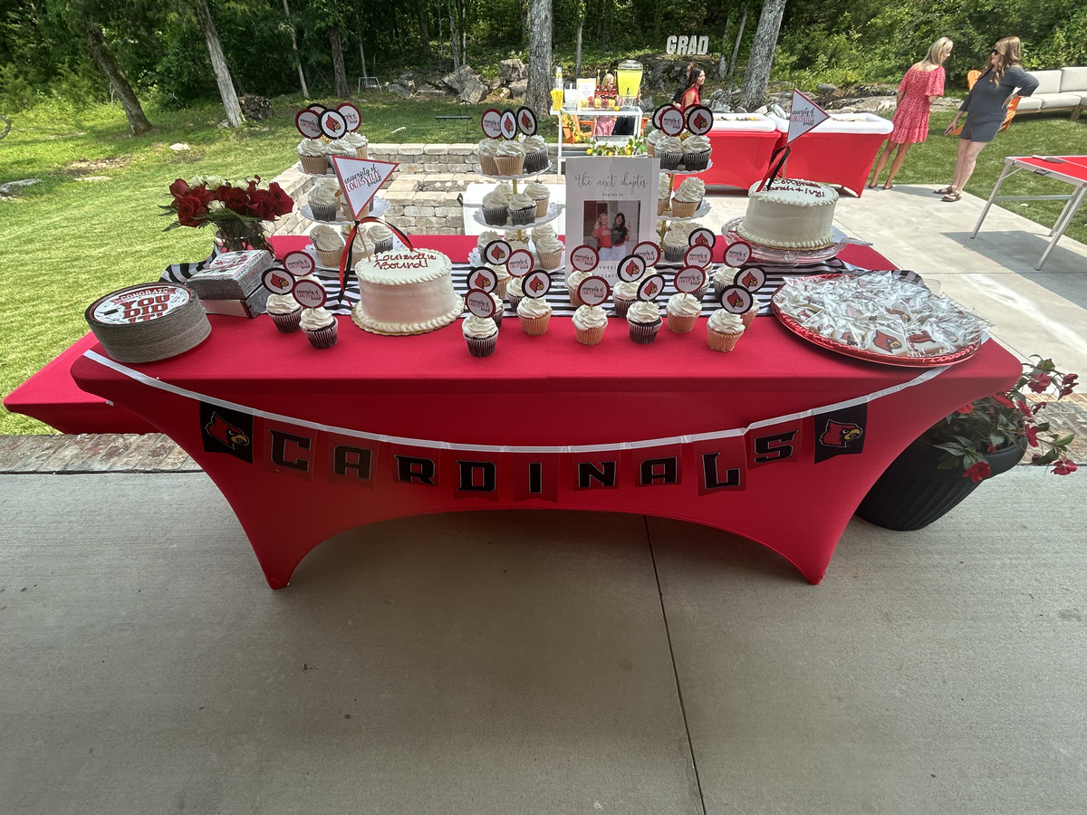 Dessert table with cupcakes topped with Louisville Cardinals toppers, two cakes, a 'Cardinals' banner draped across the front, and a framed photo of the girls