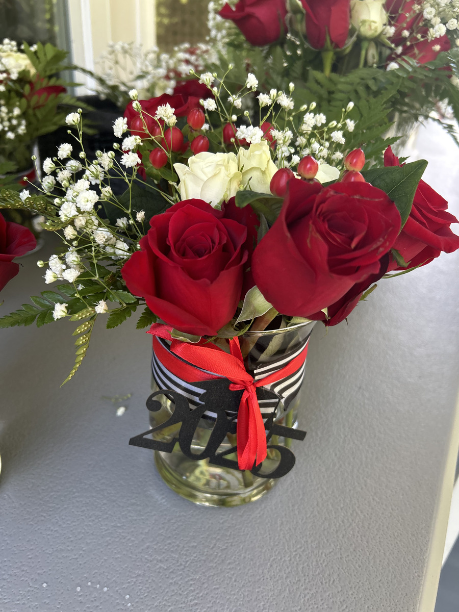 Close-up of a mason jar arrangement with red roses, white spray roses, baby's breath, hypericum berries, and a 2025 cutout on striped ribbon