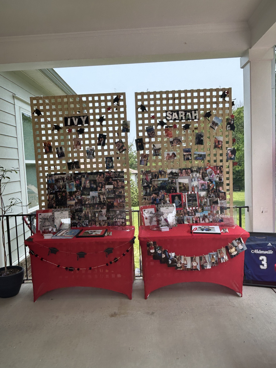 Two wood lattice panels labeled IVY and SARAH, covered in photos clipped on with mini graduation cap accents, set up behind red skirted tables