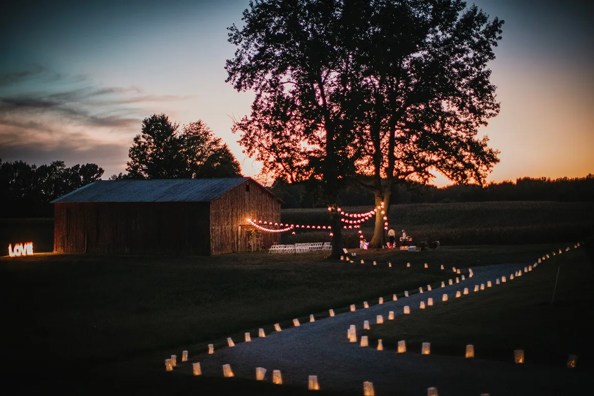 Aerial night shot of the entire venue — LOVE sign glowing on the left, luminaries lining the driveway, café lights over the reception, old barn in the background at sunset
