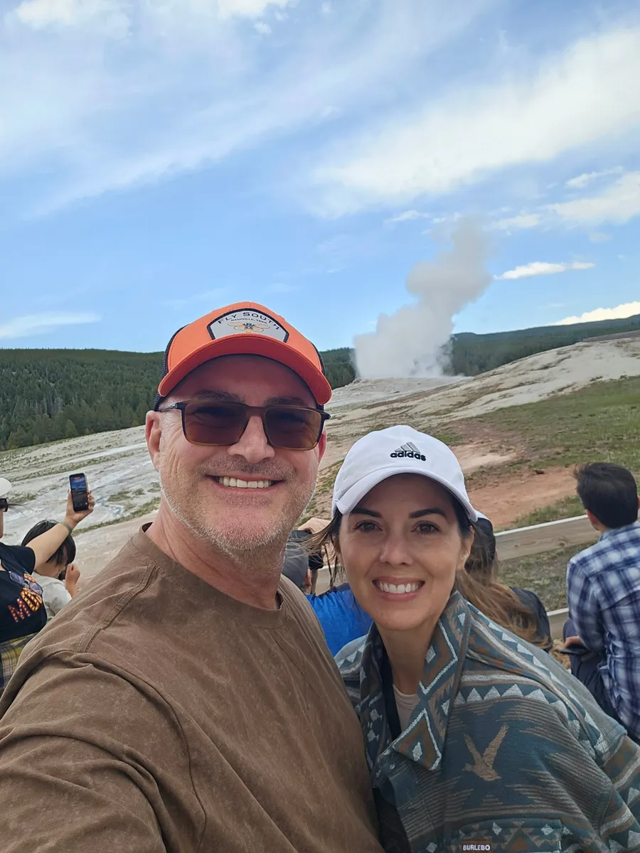Jason and Heather taking a selfie with Old Faithful erupting behind them